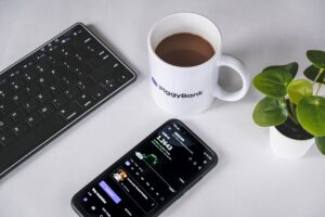 A coffee mug labeled PiggyBank, a potted plant, a wireless keyboard, and a smartphone displaying a financial app with a graph and numbers rest on the white desk—perfect for tackling that hangover while keeping an eye on your finances.