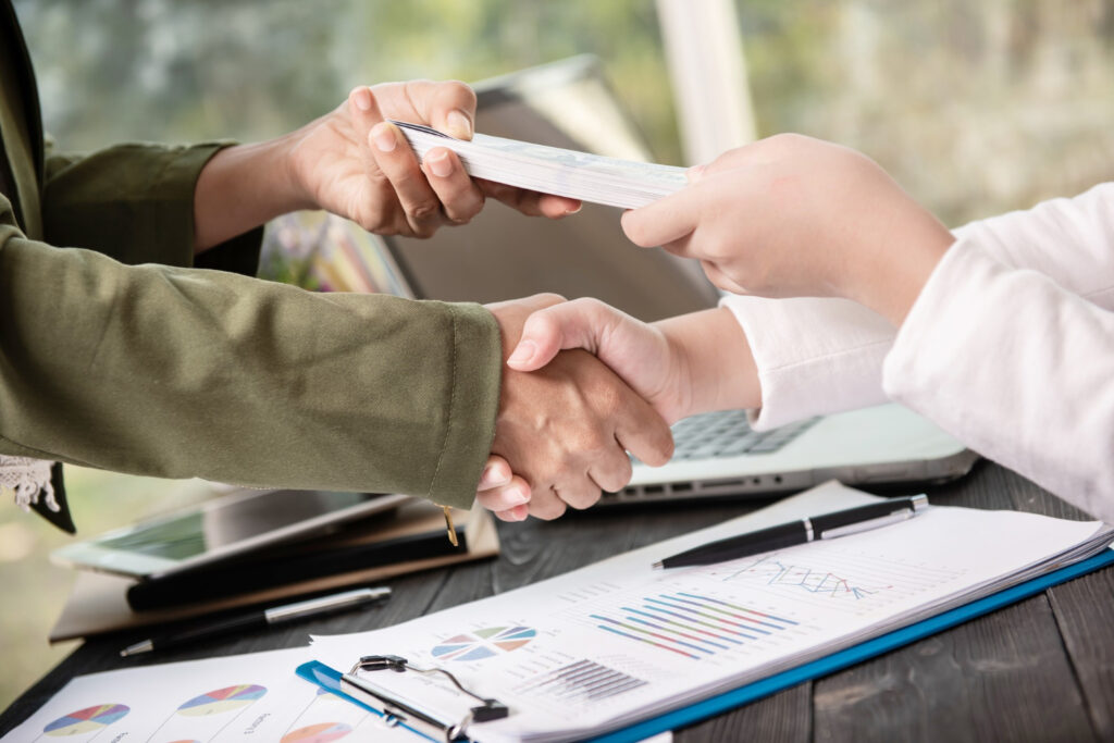 Two people shaking hands while exchanging a stack of cash over a desk with charts, graphs, and a laptop, illustrating a financial transaction such as securing small business loans for restaurants.