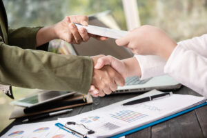 Two people shaking hands while exchanging a stack of cash over a desk with charts, graphs, and a laptop, illustrating a financial transaction such as securing small business loans for restaurants.