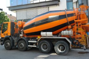 A large orange and black striped cement mixer truck, possibly funded by business loans, is parked on a city street beside a modern building with large windows.