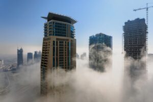 Three modern skyscrapers rise above thick morning fog, with a construction crane visible on the right. The cityscape and highways fade into the distance under a clear blue sky.