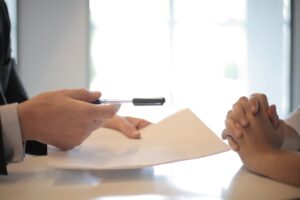 A person in a suit hands a pen and document to another person with clasped hands, suggesting a signing or agreement process at a desk in a well-lit office.