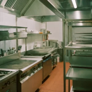 A clean, empty commercial kitchen with stainless steel appliances, shelves, and countertops. The space is well-lit, with cooking equipment neatly organized along the tiled walls and floor.