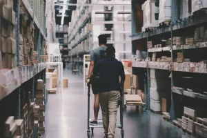 Two people move through a warehouse aisle filled with stacked boxes. One person pushes a cart while the other rides on it. The shelves around them are loaded with various packaged goods.
