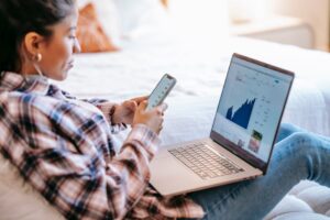 A person sitting on a bed using a smartphone, with an open laptop on their lap displaying a financial graph or stock market data.