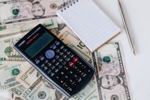 A black scientific calculator, a lined notepad, and a silver pen are placed on top of scattered U.S. dollar bills of various denominations.
