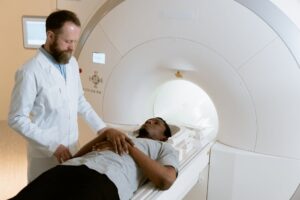 A doctor in a white coat stands beside a patient lying on a table about to enter an MRI machine, preparing for a medical scan. The patient appears calm, with hands resting on their stomach.