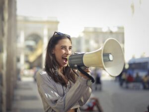 A woman standing outdoors, smiling and speaking into a large megaphone. She is wearing sunglasses on her head and a light jacket, with a blurred cityscape in the background.