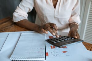 A person wearing a light shirt uses a calculator at a desk with papers, charts, and a lined notepad. Only their hands and part of their torso are visible.