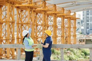 Two construction workers wearing hard hats and safety vests stand on a site, with one pointing up at large yellow scaffolding and the other holding a radio. A partially built structure is visible in the background.