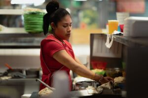 A woman wearing a red dress works in a kitchen, focused on preparing food at a counter. Plates, containers, and a drink are visible around her in the busy workspace.