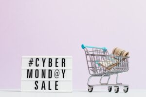 A lightbox sign with the text #CYBER MONDAY SALE sits next to a small shopping cart filled with rolled-up US dollar bills, against a light pink background.