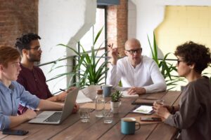 Four people sit around a wooden table in a modern office, engaged in a discussion. Laptops, tablets, notebooks, and coffee cups are on the table, with green plants and large windows in the background.