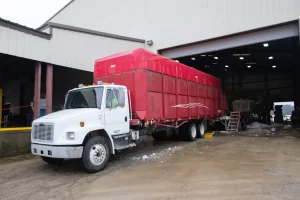 A white semi-truck with a large red trailer is parked outside a warehouse, partially inside an open garage door on a wet, muddy ground. The warehouse interior is dimly lit.