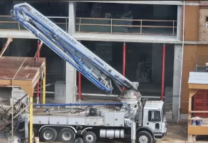 A large cement pump truck labeled AMERICAN STANDARD is parked at a construction site, with its long articulated boom extended upward toward a building under construction.