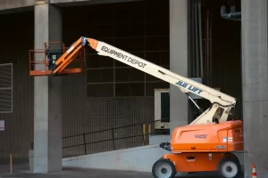 An orange JLG lift with “Equipment Depot” branding extends its arm to reach a platform near a concrete building, likely for construction or maintenance work.