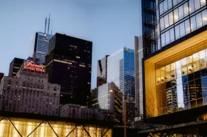 Cityscape at dusk featuring tall modern glass buildings and older skyscrapers, including the Fairmont Royal York hotel with its illuminated red sign; office lights are visible through some windows.