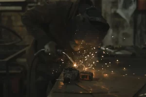 A person wearing protective gear welds metal on a workbench, creating bright sparks. The workshop environment appears dimly lit and industrial.