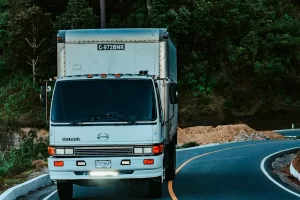 A white delivery truck with a box-shaped cargo area drives on a winding road surrounded by dense green trees and foliage.