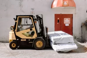 A yellow and black forklift moves two large, white-wrapped packages in front of a red door with the Home Hardware logo, set against a white building exterior.