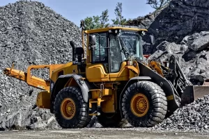 A yellow construction loader with large tires is parked on rocky ground, surrounded by piles of gravel and rocks, with a rocky hillside and sparse trees in the background.