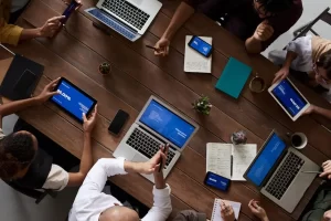 A group of people sit around a wooden table using laptops, tablets, and smartphones. Notebooks, papers, and a small plant are also on the table, suggesting a collaborative work or meeting environment.