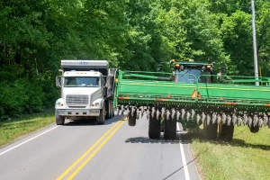A large green tractor with wide farm equipment attachment passes closely by a white dump truck on a narrow rural road bordered by green trees.