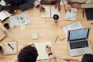 Four people sit around a wooden table with laptops, charts, notebooks, and coffee cups, actively writing and discussing documents in a collaborative work setting.