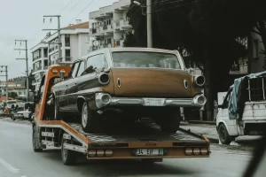 A vintage brown and beige car is loaded onto a flatbed tow truck on a city street, with buildings, trees, and other vehicles in the background.