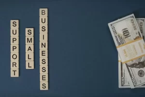 Scrabble tiles spell Support Small Businesses on a dark blue background, with a stack of $100 bills bound by a band placed to the right.