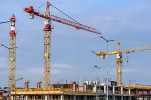 Several tall construction cranes tower over a building under construction, with concrete columns and scaffolding visible. The sky is mostly clear with a few clouds.