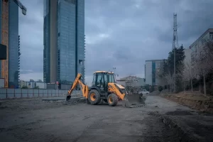 A yellow excavator is parked on a construction site with tall modern buildings and cranes in the background under a cloudy sky.