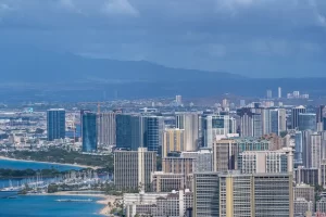 Aerial view of a coastal city with numerous high-rise buildings, a marina with boats, sandy beaches, and mountains in the background under a cloudy sky.