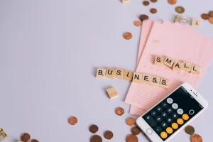 Scrabble tiles spelling SMALL BUSINESS, a smartphone with a calculator app open, pink notepads, and scattered coins on a light surface.