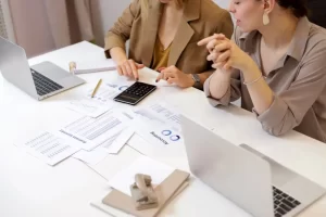 Two women sit at a desk with laptops, calculator, and business documents, reviewing charts and spreadsheets. One points at the calculator while the other listens attentively.