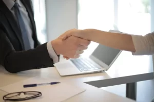 Two people shaking hands across a desk with a laptop, pen, and papers, suggesting a business or interview setting. Only their arms and hands are visible.