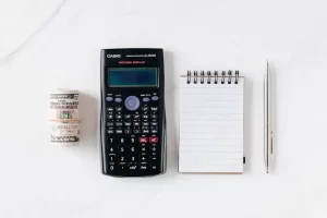 A rolled stack of U.S. dollar bills, a scientific calculator, a blank spiral notepad, and a silver pen arranged neatly on a white surface.