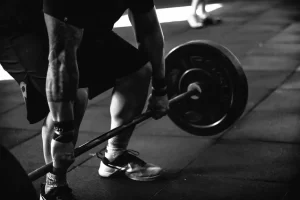 Black and white photo of a person in athletic wear gripping a barbell on a gym floor, preparing to lift weights. The focus is on their muscular arms and legs, with another person visible in the background.