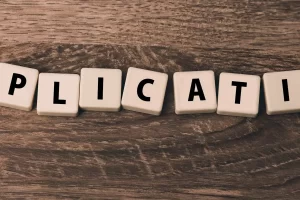 Scrabble tiles spelling out part of the word APPLICATION are arranged in a row on a wooden surface, showing the letters PLICATI.