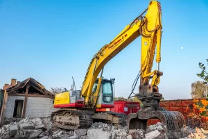 A yellow excavator with a claw attachment sits on rubble, demolishing a building with a partially collapsed roof under a clear blue sky.