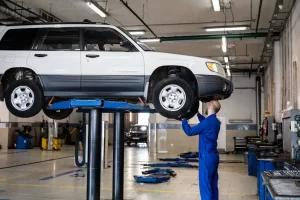A mechanic in blue coveralls inspects the underside of a white SUV raised on a hydraulic lift inside a spacious, well-lit auto repair shop.