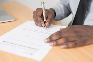 A person in a dress shirt and tie is sitting at a wooden desk, holding a pen and signing a document or contract on white paper.