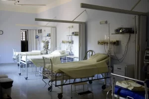 Three empty hospital beds with white linens are arranged in a clean, well-lit hospital room. Medical equipment and monitors are mounted on the walls beside each bed. The room appears calm and organized.