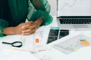 A person with folded hands sits at a desk covered with financial charts, graphs, documents, a magnifying glass, a pencil, a pen, pills in a blister pack, and a laptop showing a stock market graph.