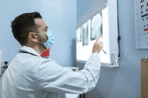 A doctor wearing a white coat and a face mask examines and points at a brain X-ray displayed on a lightbox in a medical office. An eye chart is partially visible on the wall nearby.