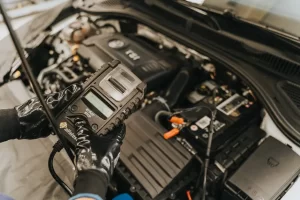A person wearing black gloves uses a diagnostic scanner on a car engine, checking for issues under the hood in a vehicle repair setting.