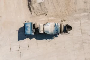 Aerial view of a blue cement mixer truck parked on a sandy construction site, with tire tracks and concrete slabs visible on the ground.