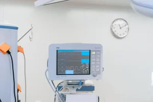 A digital medical monitor displaying vital signs sits in a clean hospital room. A wall-mounted clock in the background shows the time as 11:10. Medical equipment is visible to the side.