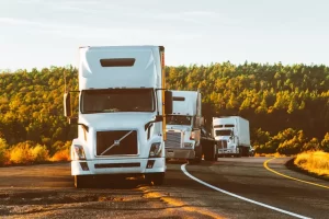 Three semi-trucks are parked on the side of a curved road, with trees and greenery in the background under a clear sky.