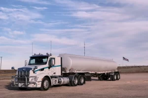A white tanker truck with a large cylindrical trailer is parked on a gravel lot under a partly cloudy sky. An American flag and some utility poles are visible in the background.